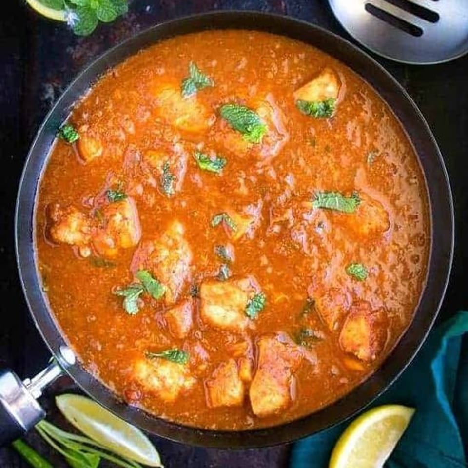 Overhead view of spicy red curry with paneer, mint garnish, lemon wedges, herbs, and a slotted spoon.