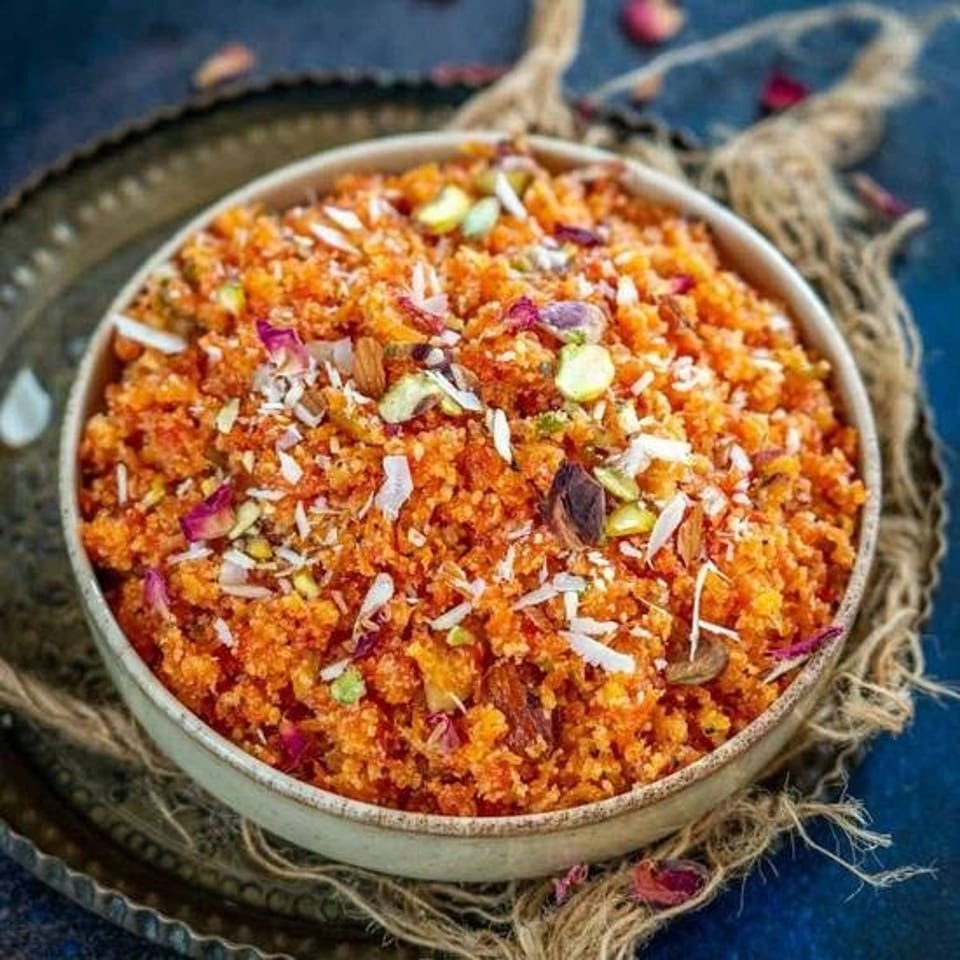 Bowl of orange crumbly dessert topped with pistachios, coconut flakes, almonds, and dried rose petals on a decorative tray.