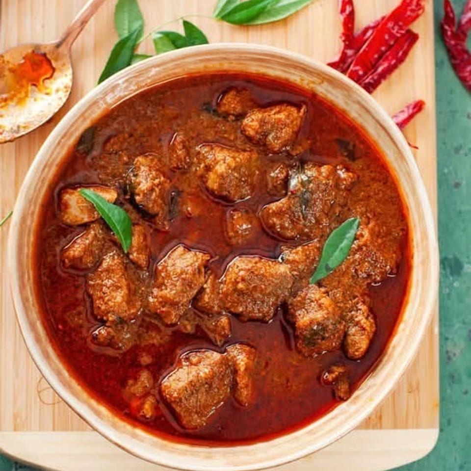 Overhead view of spicy red curry with browned meat, green herbs, dried chilies, rice, and cucumber slices on a wooden board.